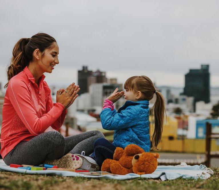 Clinician and child sit on a blanket outdoors, smiling and playing a clapping game.