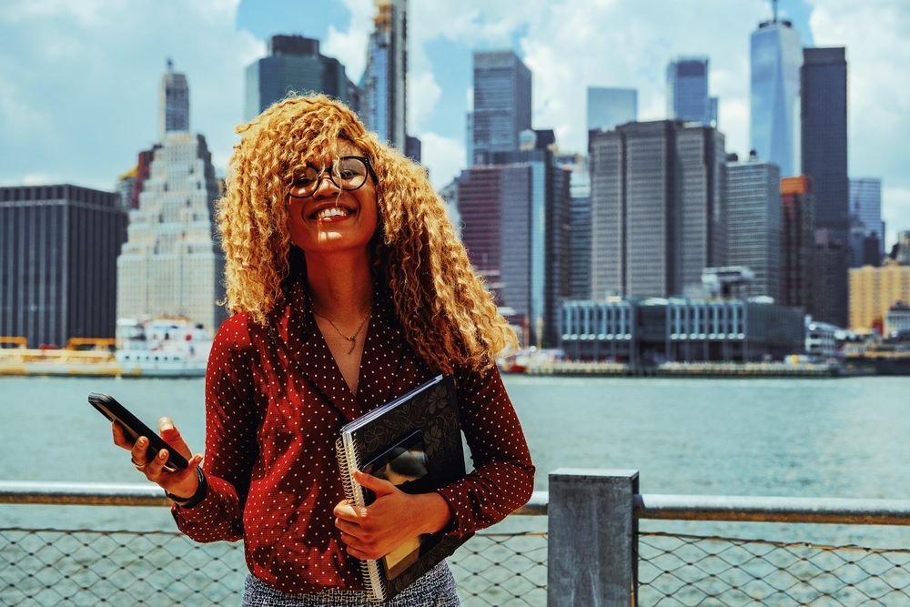 Woman looks ahead smiling happily holding a phone and a spiral notebook, a NY city skyline behind her.