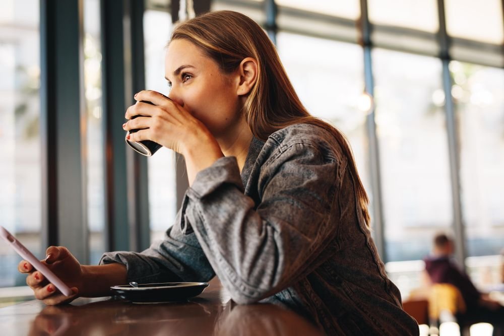 Woman sits at a coffee shop, holding a phone, taking a sip of coffee from a black porcelain mug.