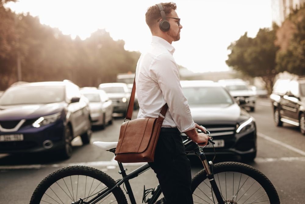 Hipster guy wearing headphones and a leather satchel, walks his bike across an intersection.