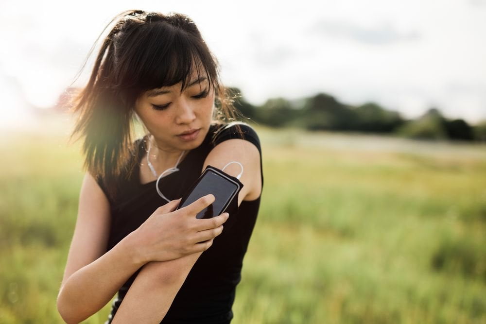 Woman outdoors in a green space, checks her iPhone, which is in a phone holder on her arm.