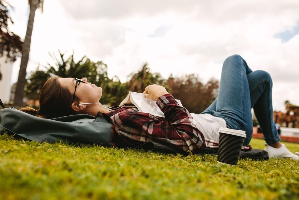 Woman lies outdoors on the grass, wearing earbuds, looking to the sky, a book on her chest, and paper coffee cup next to her.